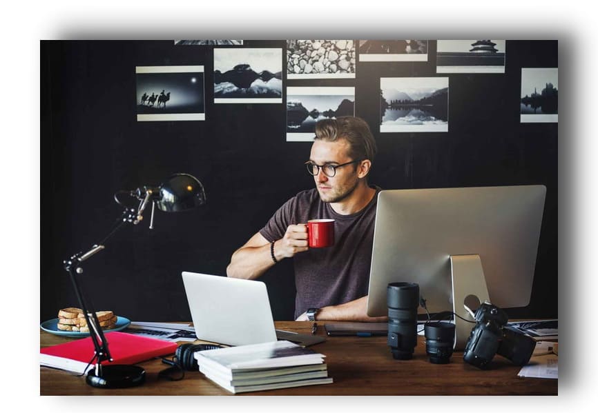 Мужчина сидит за экраном настольного компьютера и ноутбуком, держа в руках кружку кофе